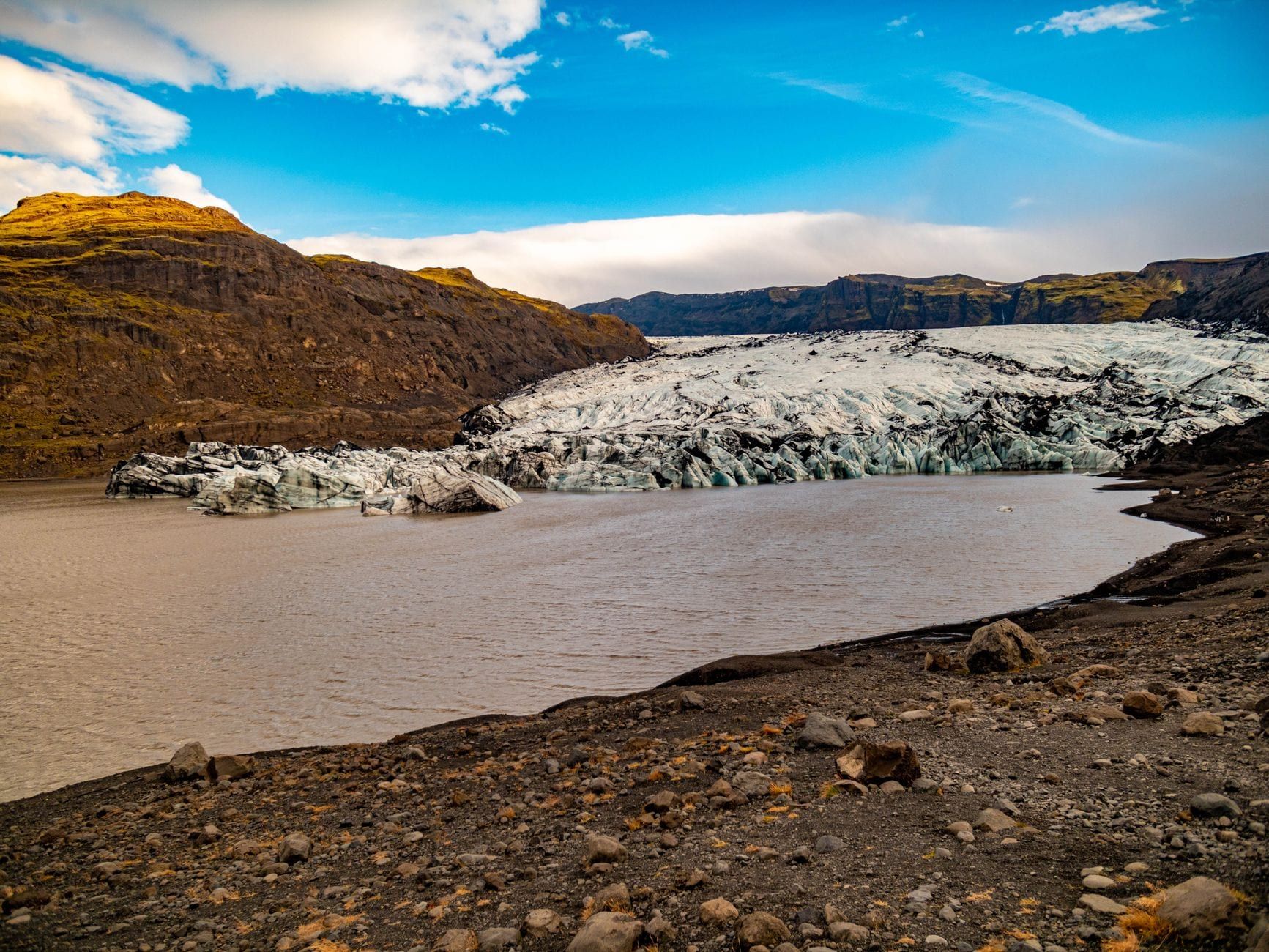 Vista amplia del glaciar Sólheimajökull, surcado por ceniza, que desemboca en una laguna glaciar junto a colinas escarpadas.