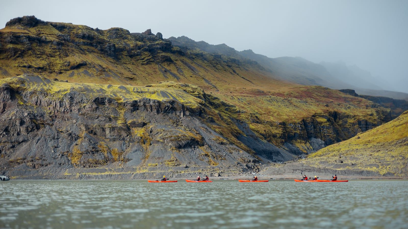 Kayakers in drysuits paddle red kayaks across the glacial lagoon in front of rugged mountain slopes at Sólheimajökull Glacier.