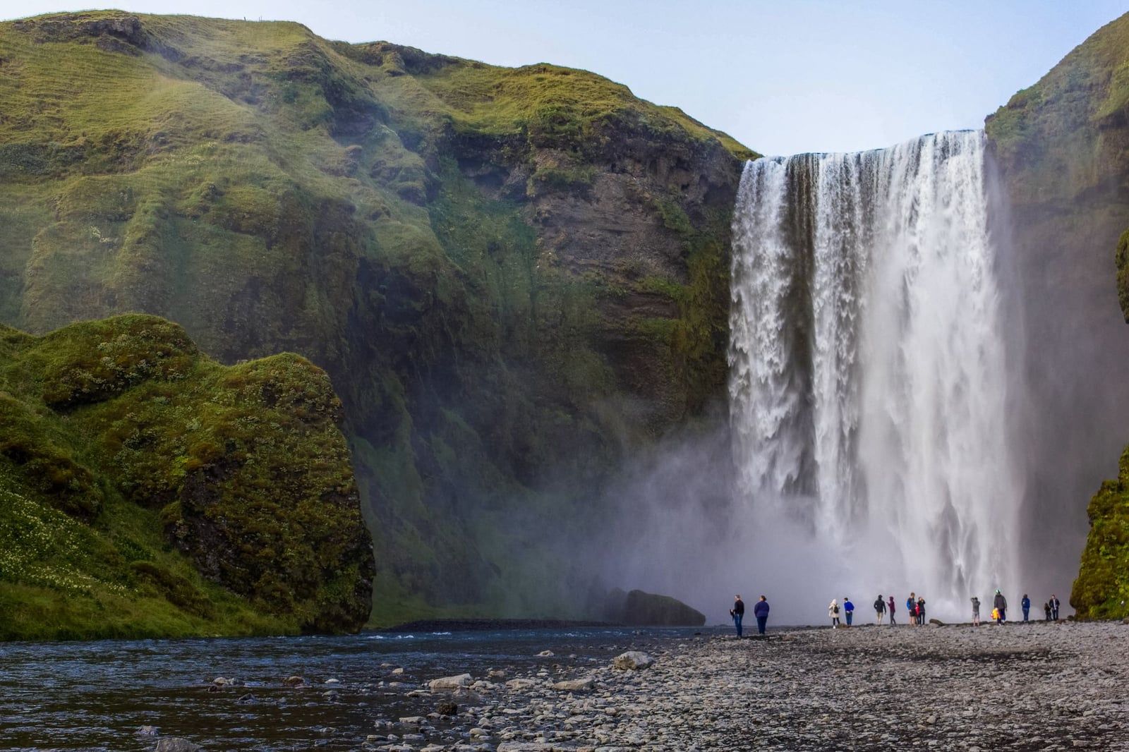 Visitors stand along the gravel riverbank watching the powerful cascade of Skógafoss waterfall near Sólheimajökull Glacier.