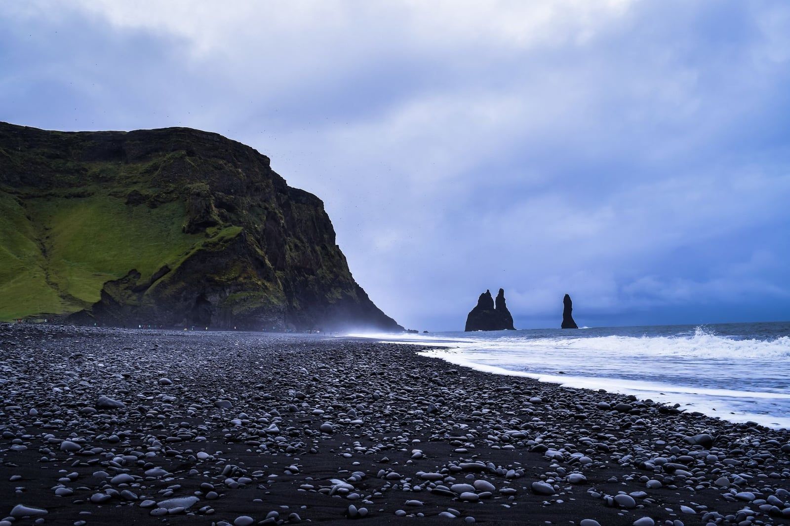 Black volcanic pebbles and sand line the coast at Reynisfjara beach, with the Reynisdrangar sea stacks visible offshore near Sólheimajökull Glacier.